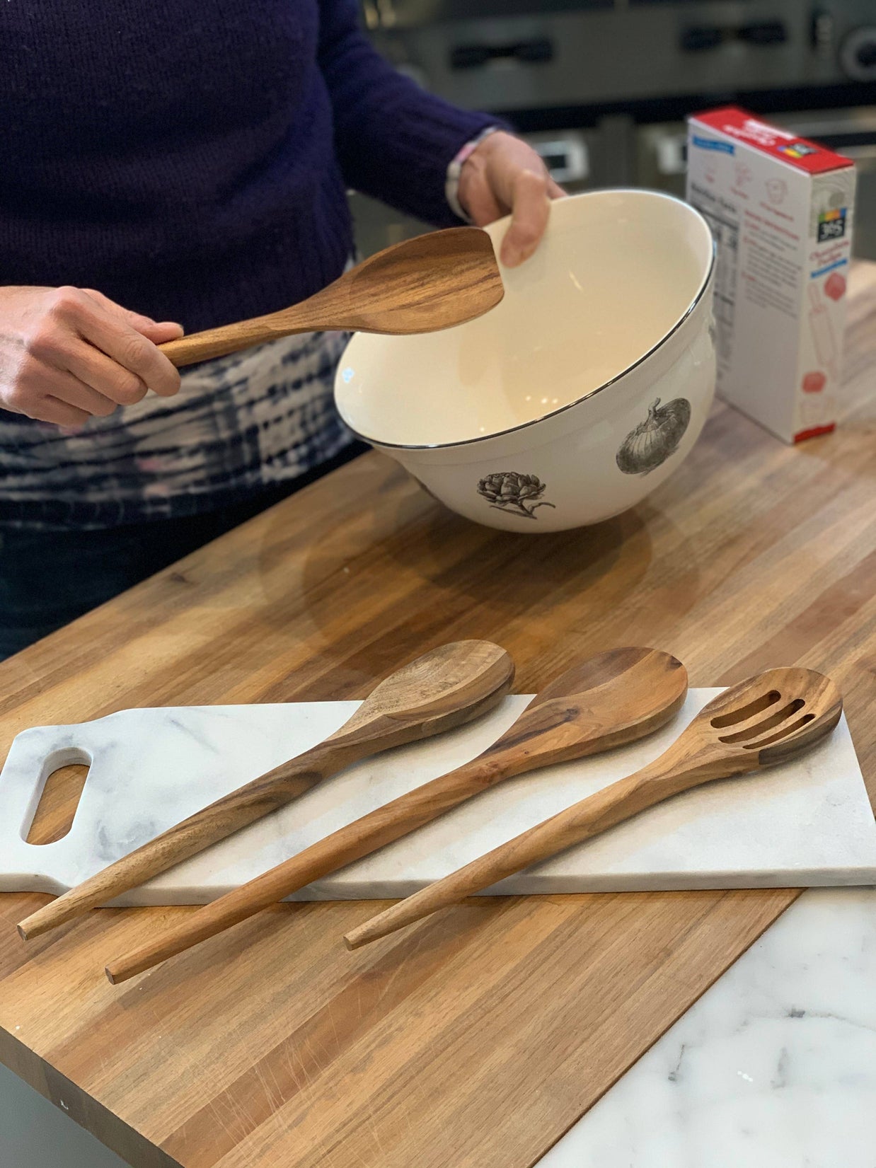 Person holding a wooden spoon over a bowl on a kitchen counter with wooden utensils in the foreground.