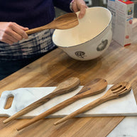Person holding a wooden spoon over a bowl on a kitchen counter with wooden utensils in the foreground.