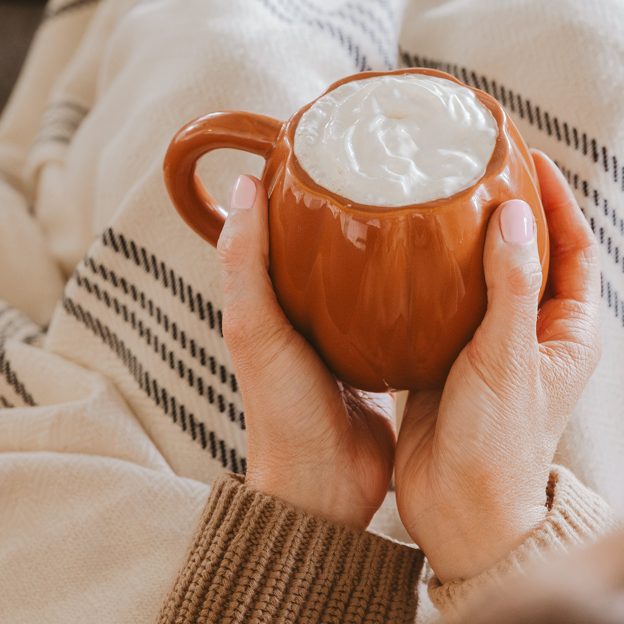 Person holding a brown mug with a hot beverage and whipped cream, wearing a beige sweater.