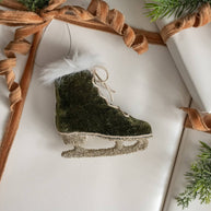 Decorative ice skate ornament with green finish and white feather on a white surface with brown ribbon.