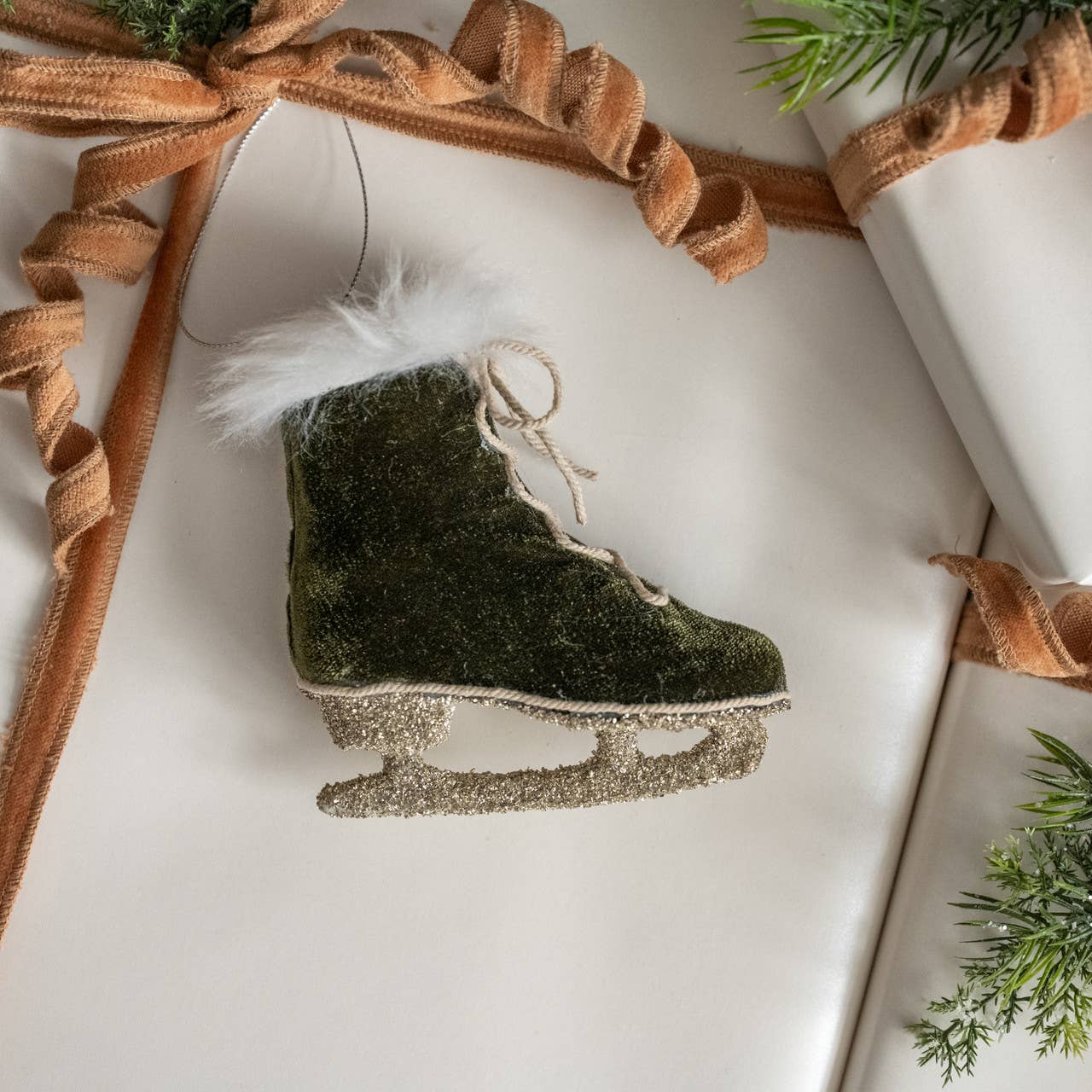 Decorative ice skate ornament with green finish and white feather on a white surface with brown ribbon.