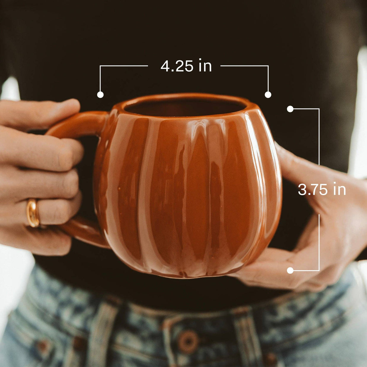 Brown ceramic mug held by hands with measurements indicated, worn person wearing a ring and denim shirt.