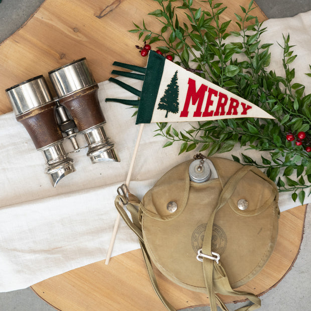 Vintage-style binoculars, leather pouch, and Christmas-themed pennant with greenery on a wooden surface.